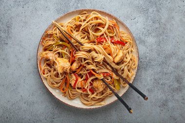 Tavuk eti, havuç, dolma biber ve diğer sebzelerle kızartılmış Udon erişteleri gri arka planda çubuklarla servis edilir ve susam tohumları serpiştirilir.