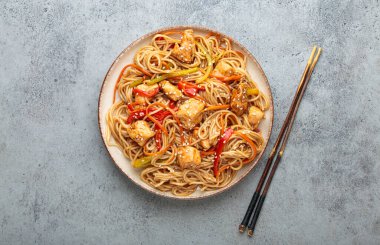 Tavuk eti, havuç, dolma biber ve diğer sebzelerle kızartılmış Udon erişteleri gri arka planda çubuklarla servis edilir ve susam tohumları serpiştirilir.