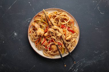 Lezzetli udon erişteleri tavuk eti ve sebzeyle kızartılmış, susam tohumları serpiştirilmiş, yemek çubuklarıyla servis edilmiş, karanlık bir arka planda, iştah açıcı bir Asya mutfağı sahnesi yaratıyor.