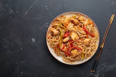 Lezzetli udon erişteleri tavuk eti ve sebzeyle kızartılmış, susam tohumları serpiştirilmiş, yemek çubuklarıyla servis edilmiş, karanlık bir arka planda, iştah açıcı bir Asya mutfağı sahnesi yaratıyor.