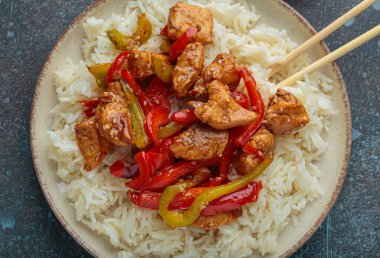 Lezzetli kızarmış tavuk, dilimlenmiş dolmalık biber ve susam tohumları. Yanında yemek çubukları olan bir kasede beyaz pirinçle servis ediliyor. Kırsal alanda Asya 'dan esinlenilmiş lezzetli bir yemek.