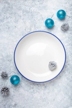 Mavi kenarlı boş beyaz tabak gümüş kozalaklar ve turkuaz noel süslemeleriyle çevrili gri desenli bir arka planda oturur ve şenlikli bir tatil masası oluşturur.