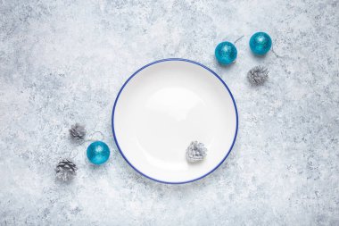Mavi kenarlı boş beyaz tabak gümüş kozalaklar ve turkuaz noel süslemeleriyle çevrili gri desenli bir arka planda oturur ve şenlikli bir tatil masası oluşturur.