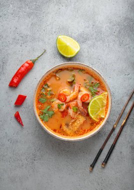 Geleneksel baharatlı Tayland çorbası Tom. İçinde karides, limon, acı biber ve kişniş var. Gri arka planda yemek çubukları ve Tayland yemeği de var.