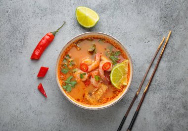 Geleneksel baharatlı Tayland çorbası Tom. İçinde karides, limon, acı biber ve kişniş var. Gri arka planda yemek çubukları ve Tayland yemeği de var.