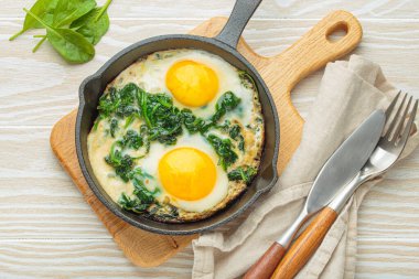 Sağlıklı ıspanaklı omlet, çırpılmış demir bir tavada iki sahanda yumurta, ahşap bir masada çatal bıçak ve peçeteyle servis edilir, besleyici bir kahvaltı veya brunch için mükemmeldir.