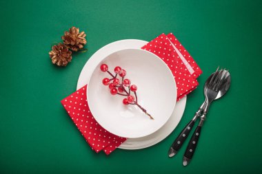 Beyaz tabak, kırmızı noktalı peçete, çatal bıçak, köknar ağacı dalı ve yeşil arka planda kutsal böğürtlen süslemesi.