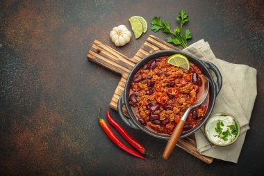 Geleneksel Meksika Chili con carne, kireç ve kırmızı biberlerle süslenmiş dökme demir bir tencerede servis edilir, ekşi krema ve taze malzemelerin yanında, kırsal bir ahşap tahta üzerinde, kopyalama alanı.