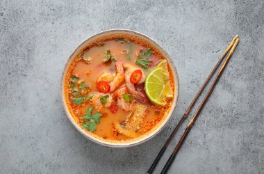 Geleneksel baharatlı Tayland çorbası Tom. İçinde karides, limon, acı biber ve kişniş var. Gri arka planda yemek çubukları ve Tayland yemeği de var.