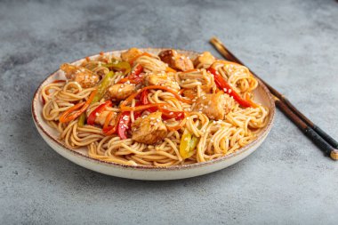 Teriyaki tavuklu erişte ve havuç da dahil olmak üzere taze sebze, susam tohumlu dolmalık erişte gri desenli arka planda servis edilir.