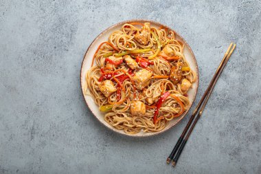 Tavuk eti, havuç, dolma biber ve diğer sebzelerle kızartılmış Udon erişteleri gri arka planda çubuklarla servis edilir ve susam tohumları serpiştirilir.