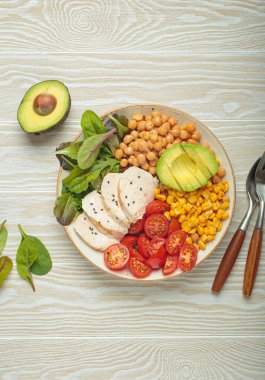 Kızarmış tavuk göğsü, avokado, nohut, mısır, domates ve karışık yeşillikler, yukarıdan beyaz ahşap bir masa, dengeli ve besleyici bir yemek.