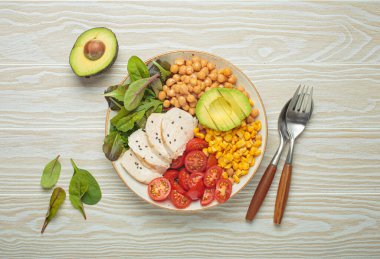 Kızarmış tavuk göğsü, avokado, nohut, mısır, domates ve karışık yeşillikler, yukarıdan beyaz ahşap bir masa, dengeli ve besleyici bir yemek.