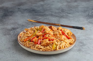 Teriyaki tavuklu waok noodle ve havuç dahil taze sebze, susam tohumlu dolma biber gri dokulu arka planda servis edilir.
