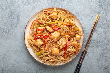 Tavuk eti, havuç, dolma biber ve diğer sebzelerle kızartılmış Udon erişteleri gri arka planda çubuklarla servis edilir ve susam tohumları serpiştirilir.