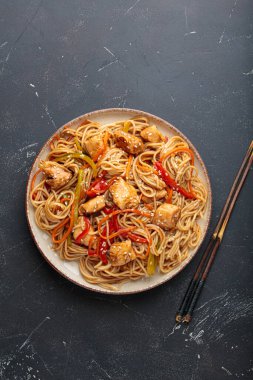 Lezzetli udon erişteleri tavuk eti ve sebzeyle kızartılmış, susam tohumları serpiştirilmiş, yemek çubuklarıyla servis edilmiş, karanlık bir arka planda, iştah açıcı bir Asya mutfağı sahnesi yaratıyor.