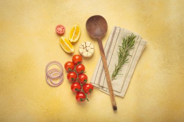 Kiraz domatesleri, sarımsak, limon dilimleri, havuç, kırmızı soğan ve biberiye filizi sarı bir zemin üzerinde ahşap kaşık, aşçılık yaratıcılığı ve sağlıklı beslenme konsepti ile birlikte sıralanmış.