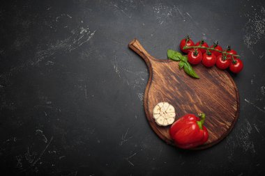 Kiraz domatesleri, kırmızı dolma biber ve taze fesleğen yaprakları ahşap bir kereste kesme tahtasının yanında koyu dokulu bir arka planda duruyor.