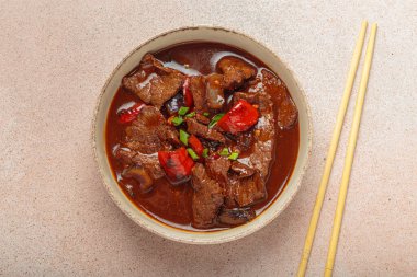 Kırmızı çan biberi, soğan ve yeşil soğanlı Asya sığır eti güveci, bir kâsede servis edilir, açık bej arka planda bir çift çubuğun yanında yer alır.