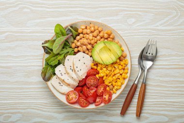 Kızarmış tavuk göğsü, avokado, nohut, mısır, domates ve karışık yeşillikler, yukarıdan beyaz ahşap bir masa, dengeli ve besleyici bir yemek.