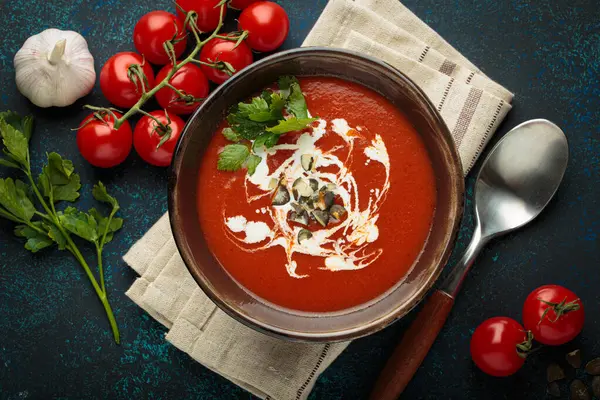 Taze domates çorbası krema, tohum ve otlarla dolu bir kasede servis edilir, yanında da mavi bir zemin üzerinde kaşık ve peçeteyle kiraz domatesleri, sarımsak ve maydanoz bulunur.