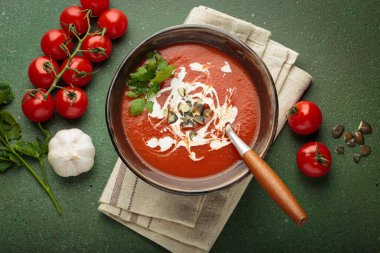 Taze domates çorbası krema, tohum ve otlarla dolu bir kasede servis edilir, yanında da yeşil bir zemin üzerinde yeşil bir kaşık ve peçeteyle kiraz domates, sarımsak ve maydanoz bulunur.