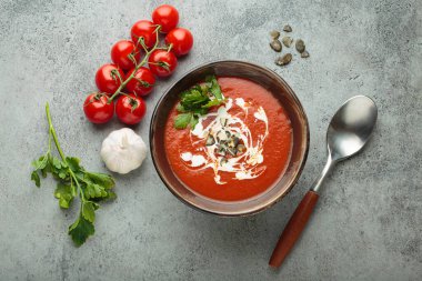 Krema, tohum ve otlarla dolu bir kasede taze domates çorbası servis edilir, yanında da kaşık ve peçeteyle birlikte kiraz domatesleri, sarımsak ve maydanoz bulunur.