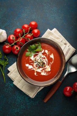 Taze domates çorbası krema, tohum ve otlarla dolu bir kasede servis edilir, yanında da mavi bir zemin üzerinde kaşık ve peçeteyle kiraz domatesleri, sarımsak ve maydanoz bulunur.