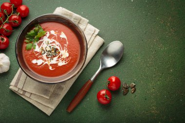 Taze domates çorbası krema, tohum ve otlarla dolu bir kasede servis edilir. Yanında da yeşil bir zemin üzerinde yeşil bir kaşık ve peçeteyle domates, sarımsak ve maydanoz bulunur.