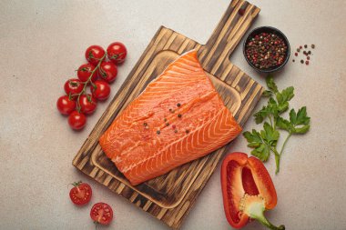 Taze somon fileto, ahşap kesim tahtasının üzerinde biber, kiraz domatesleri, kırmızı dolmalık biber ve maydanozla birlikte. Bej arka planda sağlıklı yemek malzemeleri. Üst görünüm bileşimi