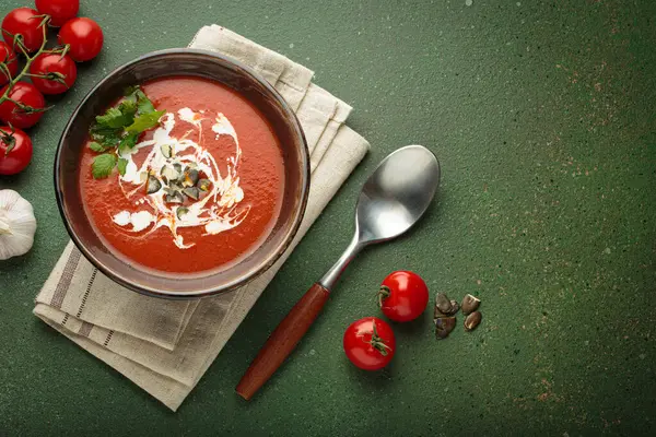 Taze domates çorbası krema, tohum ve otlarla dolu bir kasede servis edilir. Yanında da yeşil bir zemin üzerinde yeşil bir kaşık ve peçeteyle domates, sarımsak ve maydanoz bulunur.