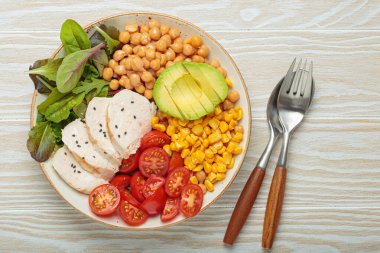 Kızarmış tavuk göğsü, avokado, nohut, mısır, domates ve karışık yeşillikler, yukarıdan beyaz ahşap bir masa, dengeli ve besleyici bir yemek.