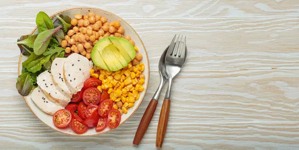 Kızarmış tavuk göğsü, avokado, nohut, mısır, domates ve karışık yeşillikler, yukarıdan beyaz ahşap bir masa, dengeli ve besleyici bir yemek.