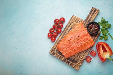 Taze somon fileto, ahşap kesim tahtasının üzerinde biber, kiraz domatesleri, kırmızı dolmalık biber ve maydanozla birlikte. Mavi arka planda sağlıklı yemek malzemeleri. Üst görünüm, alanı kopyala