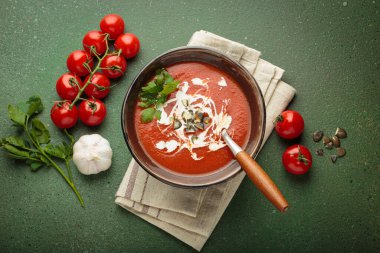 Taze domates çorbası krema, tohum ve otlarla dolu bir kasede servis edilir, yanında da yeşil bir zemin üzerinde yeşil bir kaşık ve peçeteyle kiraz domates, sarımsak ve maydanoz bulunur.