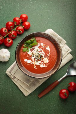 Taze domates çorbası krema, tohum ve otlarla dolu bir kasede servis edilir, yanında da yeşil bir zemin üzerinde yeşil bir kaşık ve peçeteyle kiraz domates, sarımsak ve maydanoz bulunur.