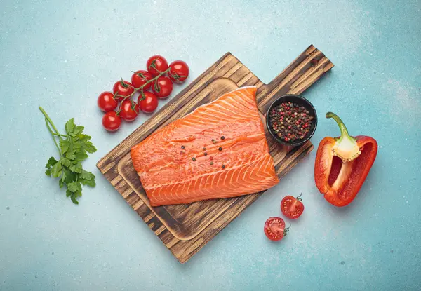 Taze somon fileto, ahşap kesim tahtasının üzerinde biber, kiraz domatesleri, kırmızı dolmalık biber ve maydanozla birlikte. Mavi arka planda sağlıklı yemek malzemeleri. Üst görünüm bileşimi