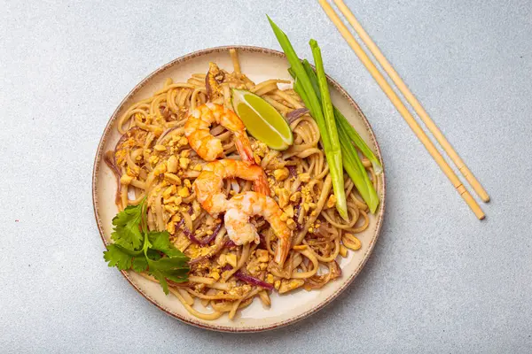 Tayland yemeği, Tayland eriştesi, karides, fıstık, limon ve yeşil soğanlı. Tabak gri bir arka planda ve yanında çubuklar var.