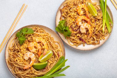 İki kase Tayland yemeği, karides, fıstık, limon ve yeşil soğanlı Tayland eriştesi. Tabak gri bir arka planda ve yanında çubuklar var.