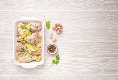 Çiğ tavuk budu, baharatlar, otlar, beyaz seramik güveçte limon açık ahşap arka planda üst manzara. Terbiye edilmiş tavuk budu ile sağlıklı yemek hazırlıyorum. Metin için boşluk.