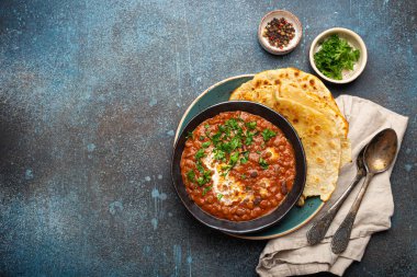 Geleneksel Hint Punjabi yemeği Dal Makhani mercimek ve siyah kasede fasulye ile birlikte naan düz ekmek, taze kişniş ve iki kaşık mavi beton kırsal masa manzaralı. Metin için boşluk.
