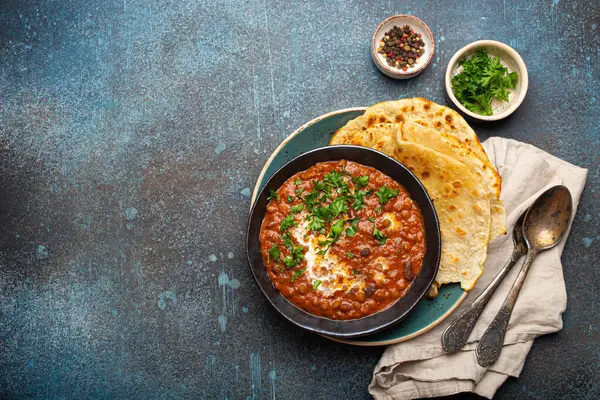 Geleneksel Hint Punjabi yemeği Dal Makhani mercimek ve siyah kasede fasulye ile birlikte naan düz ekmek, taze kişniş ve iki kaşık mavi beton kırsal masa manzaralı. Metin için boşluk.