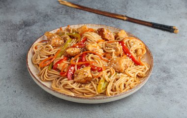 Teriyaki tavuklu waok noodle ve havuç dahil taze sebze, susam tohumlu dolma biber gri dokulu arka planda servis edilir.