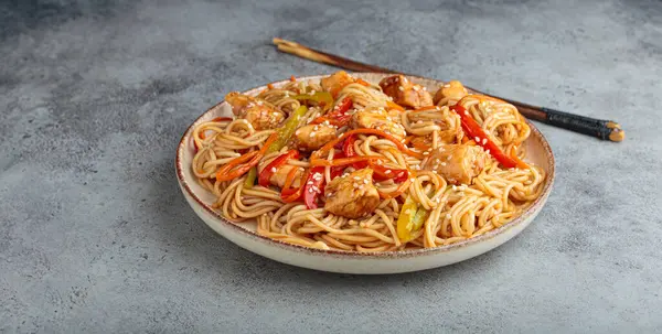 Teriyaki tavuklu waok noodle ve havuç dahil taze sebze, susam tohumlu dolma biber gri dokulu arka planda servis edilir.