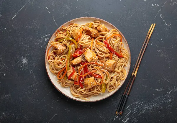 Lezzetli udon erişteleri tavuk eti ve sebzeyle kızartılmış, susam tohumları serpiştirilmiş, yemek çubuklarıyla servis edilmiş, karanlık bir arka planda, iştah açıcı bir Asya mutfağı sahnesi yaratıyor.