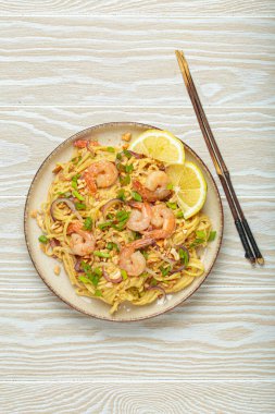 Karides, fıstık, yeşil soğan, limon dilimleri ile süslenmiş Tayland erişteleri ve beyaz ahşap bir masada servis edilen çubuklarla Tayland mutfağı.