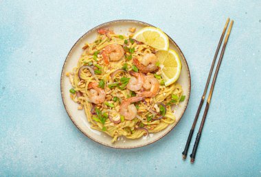 Karides, fıstık, yeşil soğan, limon dilimleri ile süslenmiş Tayland erişteleri ve açık mavi arka planda servis edilen çubuklarla Tayland mutfağı.