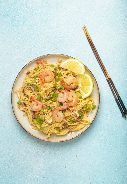 Karides, fıstık, yeşil soğan, limon dilimleri ile süslenmiş Tayland erişteleri ve açık mavi arka planda servis edilen çubuklarla Tayland mutfağı.