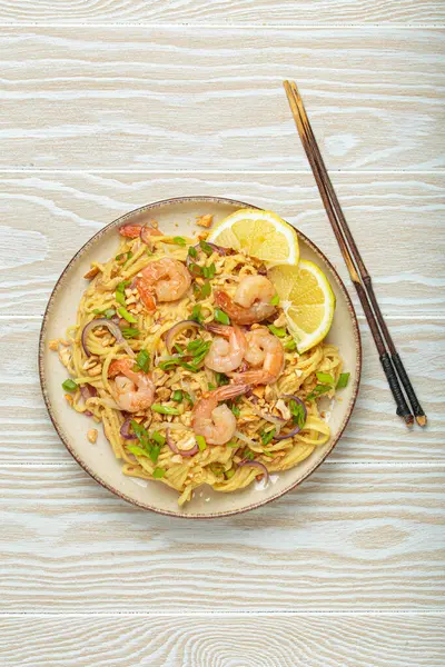 Karides, fıstık, yeşil soğan, limon dilimleri ile süslenmiş Tayland erişteleri ve beyaz ahşap bir masada servis edilen çubuklarla Tayland mutfağı.