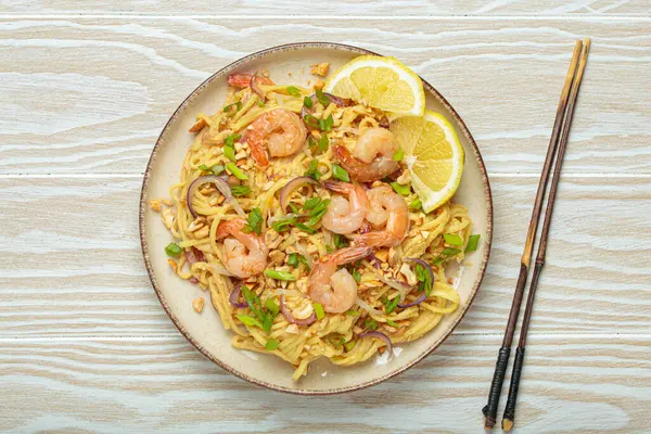 Karides, fıstık, yeşil soğan, limon dilimleri ile süslenmiş Tayland erişteleri ve beyaz ahşap bir masada servis edilen çubuklarla Tayland mutfağı.